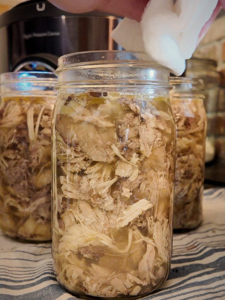 Hand wiping the rim of a quart jar of chicken and broth with a paper towel